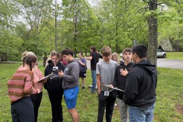 Students stand outdoors in small groups with clip boards