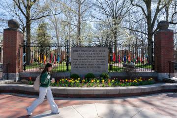 An OHIO student walks in front of the College  Gate, which has colorful flowers in front of it and colorful trees behind it, on a spring day