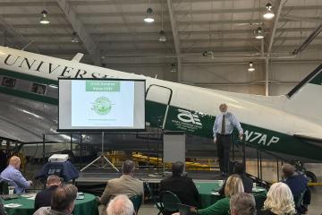 A speaker gives a presentation in an airplane hangar while standing on a stage in front of an Ohio University plane