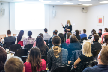 speaker at front of the room looking at presentation in front of full audience
