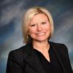 Julie Bolen Headshot. Caucasian woman, with chin length blonde hair smiling at the camera. She is wearing a black shirt and the photo backdrop is textured in gray and blues. 