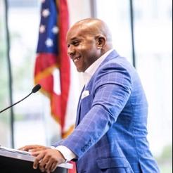 Headshot of Gary Allen, African-American man speaking at a podium in a blue suit with white shirt with an American flag in the background