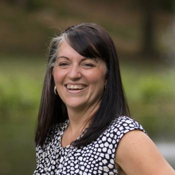 Headshot of Erin Gibson. Woman smiling towards camera with shoulder length dark hair, wearing a printed top. The background is outdoors with grass and trees behind her. 