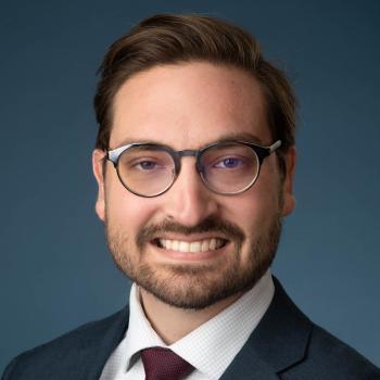 Headshot of Samuel Jurgena. A man smiles at the camera with short brown hair and a short brown beard. He is wearing dark framed glasses, a dark suit jacket, white collared shirt and a dark burgundy tie. The backdrop is a textured blue pattern. 