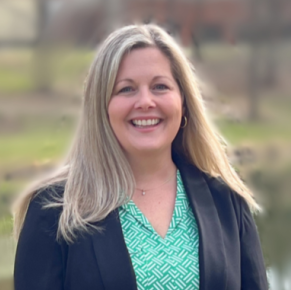 Headshot of Brooke Enderle. Woman smiles at the camera with long blonde hair, and is wearing a green blouse underneath a navy blazer. She is standing outdoors with a backdrop of trees and grass.