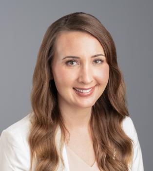 A professional headshot of Sara Condrac. A caucasian woman smiles at the camera, she has long brown hair and is wearing a light colored suit jacket and blouse. She is infront of a backdrop that is lightly textured gray.  