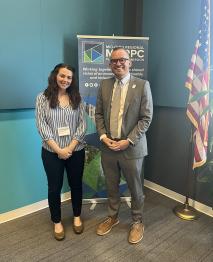 Smiling brunette woman in gray shirt and black dress pants and white name badge stands next smiling short hair man with dark glasses, gray suit and light tie. They are infront of a MORPC banner and an American flag