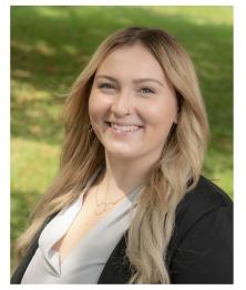 Headshot of smiling woman wearing a gray shirt and dark suit jacket with an grassy outdoor background 